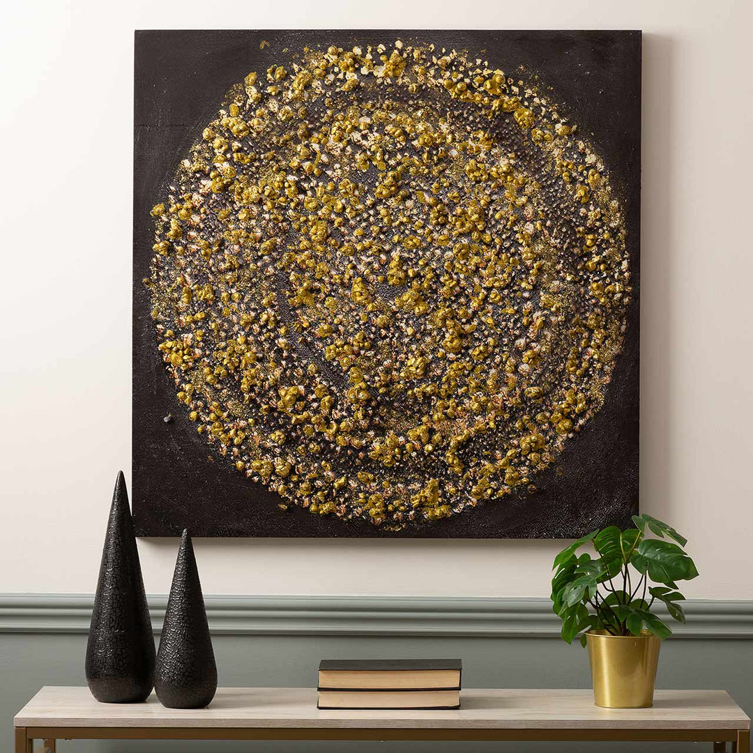 Decorative wall art with gold and black pattern on a plain wall, with two black sculptures and a plant on a table below.