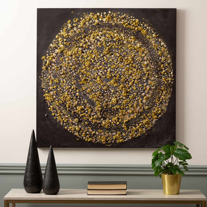 Decorative wall art with gold and black pattern on a plain wall, with two black sculptures and a plant on a table below.